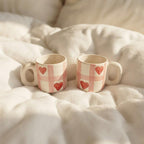 Two small ceramic mugs with cream handles and bases, decorated with a pink gingham pattern and red hearts, resting on a soft white blanket.