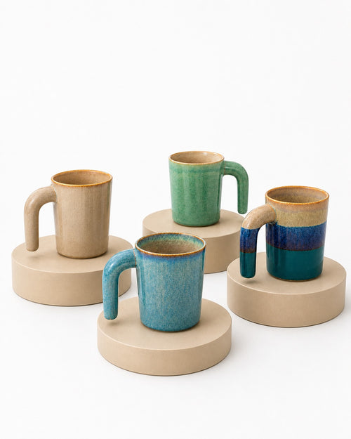 Four handmade ceramic mugs with unique handle designs, displayed on round beige pedestals against a clean white background.