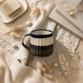 Ceramic mug with black and white design on a textured surface with dried plants and a book.