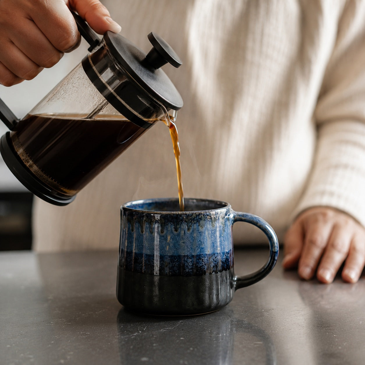 Person pouring coffee from a French press into a blue mug on a table.