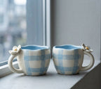 Two ceramic mugs with checkered pattern and floral handles on a windowsill.
