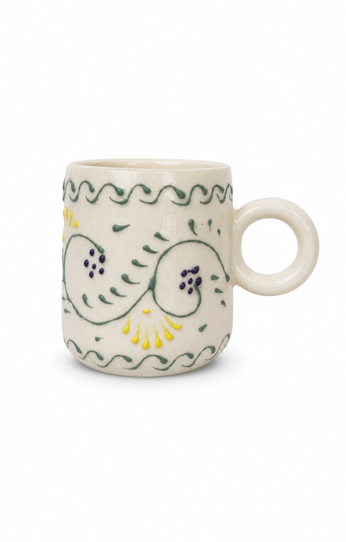 An off-white ceramic mug with a loop handle, decorated with hand-painted green and yellow floral and wavy patterns, set against a plain white background.