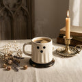 White ghost-shaped mug with steam, candle, and dried flowers on a table.