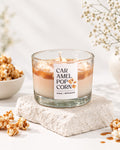 A glass jar candle labeled 'CARAMEL POP CORN' sits on a textured stone slab, surrounded by caramel popcorn pieces and dried flowers in a bright, clean setting.