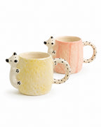 Two ceramic mugs shaped like hedgehogs, one in pale yellow and one in soft pink, both with black-spotted handles and white hedgehog heads, shown on a white background.