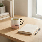 Cup with black handle, notebook, and pen on a wooden desk near a window.