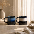 Two ceramic mugs with blue and black stripes on a stone surface with a soft background.
