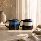 Two ceramic mugs with blue and black stripes on a stone surface with a soft background.