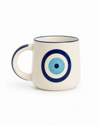 An off-white ceramic mug with a navy blue rim and handle, featuring a blue and light blue concentric circle evil eye design on the front.