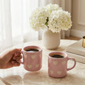 Two pink mugs with heart patterns on a marble surface, one held by a hand, with a vase of white flowers in the background.
