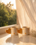 Three ceramic mugs with heart designs on a windowsill with a view of trees.