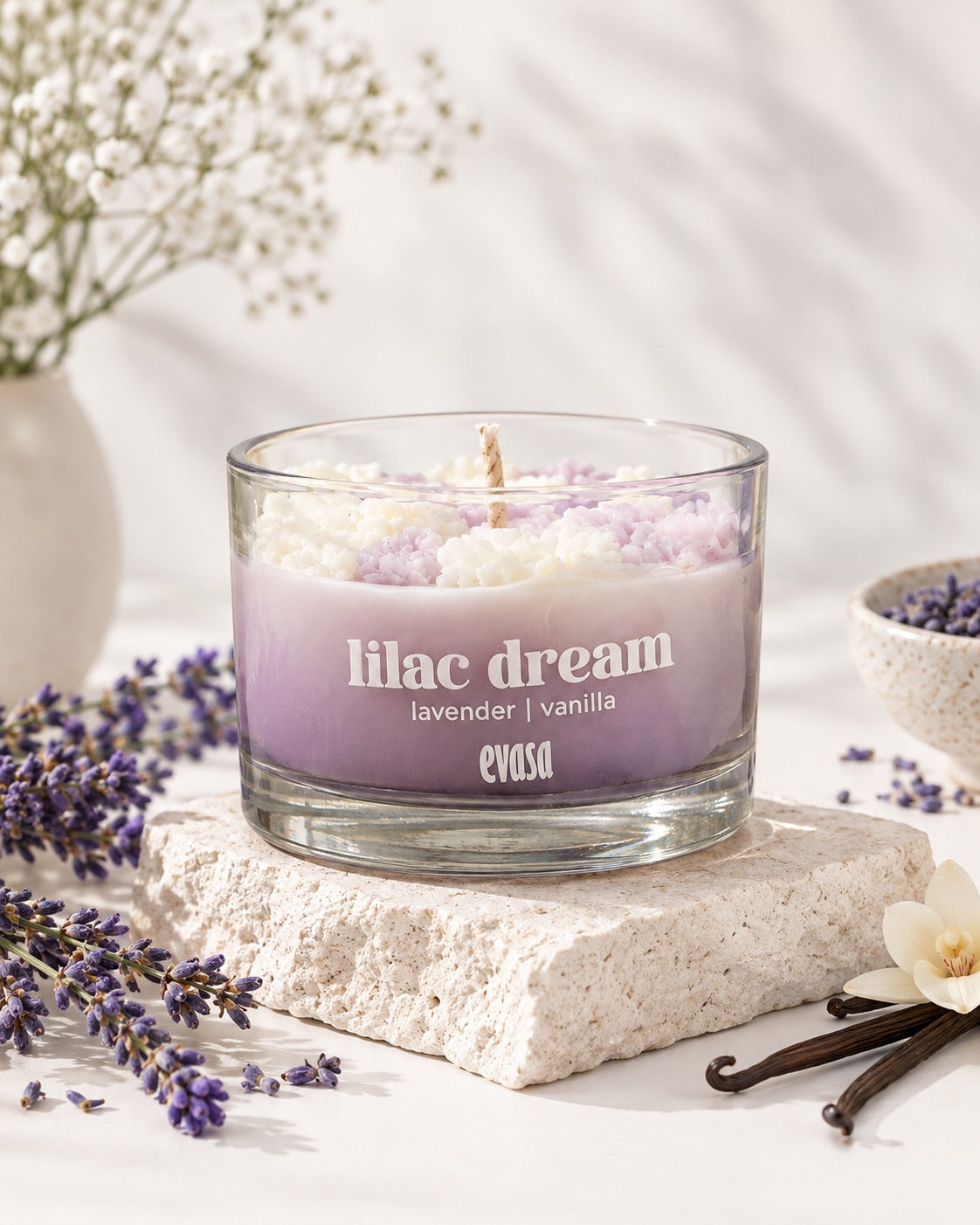 A lavender and vanilla scented candle in a clear glass jar sits on a textured stone slab, surrounded by dried lavender sprigs, vanilla beans, and small white flowers. The candle wax is swirled in light purple and white, with the text 'lilac dream lavender | vanilla' and the brand 'evasa' printed on the front.