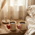 Two steaming coffee cups on a wooden table next to a bed with soft lighting.