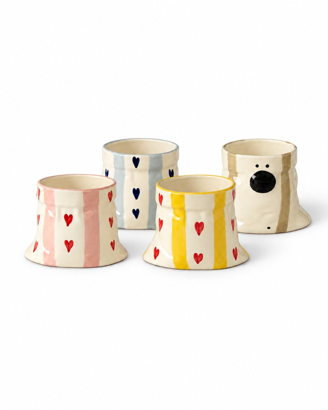 Four ceramic dog-shaped mugs on a white background. One mug features pink and cream vertical stripes with small red hearts, another has blue and cream vertical stripes with dark blue hearts, a third has yellow and cream vertical stripes with red hearts, and the fourth is cream with a black dog face and tan ears.