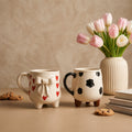Two ceramic mugs with decorative designs, a vase of tulips, and cookies on a beige surface.