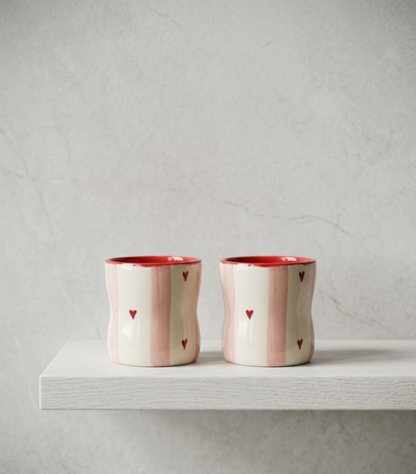 Two ceramic cups with red rims and heart patterns on a light gray surface.