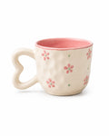 A ceramic mug with a heart-shaped handle, featuring a textured cream exterior decorated with pink flowers and small green dots, and a solid pink interior, set against a plain white background.