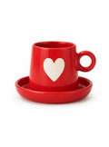 A red ceramic coffee cup with a matching saucer, featuring a white heart design on the front, set against a plain white background.