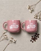 Two pink mugs with 'The Boss' text on a textured surface with flowers and coffee beans.