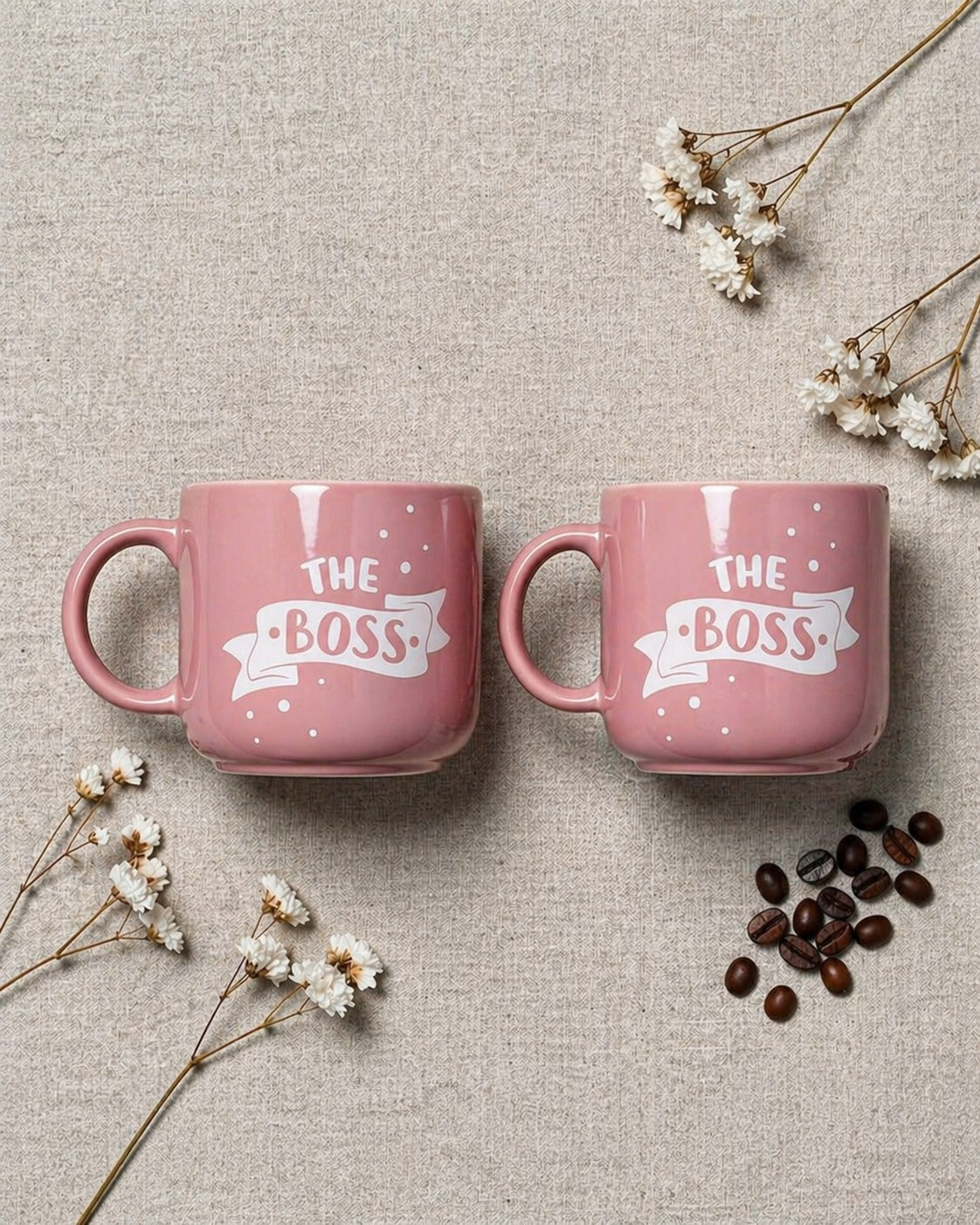 Two pink mugs with 'The Boss' text on a textured surface with flowers and coffee beans.