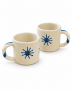 Two cream-colored ceramic mugs with blue painted starburst designs and blue handle accents, shown on a clean white background.