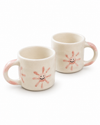 Two cream-colored ceramic mugs with pink hand-painted sunflower designs and smiling faces, set against a plain white background.