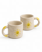 Two beige ceramic mugs with yellow smiley-face sun motifs and yellow handle accents, photographed on a clean white background.